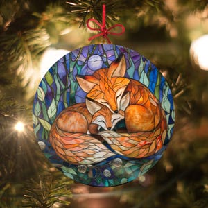 May include: Round Christmas ornament featuring two orange foxes in a stained-glass style design. The foxes are nestled together, with a full moon in the background. The ornament has a red ribbon for hanging.
