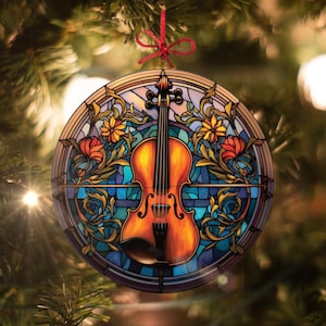 May include: A round Christmas ornament featuring a violin design. The ornament has a stained-glass effect with a brown violin at the center, surrounded by floral and leaf patterns in shades of yellow, orange, and blue. A red ribbon is attached for hanging.