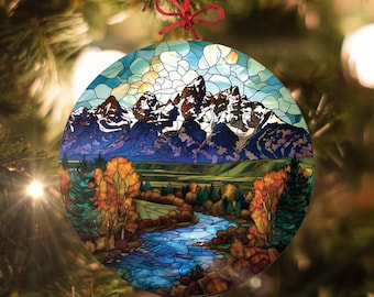May include: A round glass ornament with a stained glass design of a mountain range with a river flowing through a valley. The mountains are blue and purple with snow-capped peaks. The valley is green with trees in shades of orange, yellow, and green. The river is blue and turquoise.
