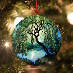 May include: Round Christmas ornament featuring a stained glass style image of a weeping willow tree over a pond. The tree has a dark brown trunk and green leaves. The pond is blue with a reflection of the sky.