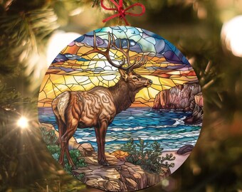 Könnte beinhalten: Ein Weihnachtsbaumschmuck aus Buntglas mit einer detaillierten Illustration eines Elchs, der auf einem felsigen Ufer mit Sonnenuntergang und Meer im Hintergrund steht.