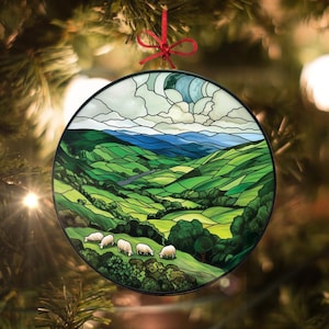 May include: Round Christmas ornament featuring a landscape scene with rolling green hills, a river, and sheep grazing. The top of the ornament depicts a cloudy sky. The ornament is trimmed with a black border and has a red ribbon for hanging.