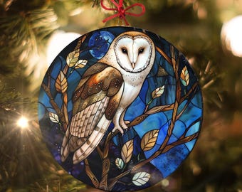 May include: A round glass ornament with a stained glass design of a barn owl perched on a branch with blue and brown leaves. The owl has white feathers with brown markings and yellow eyes. The background is a dark blue with a full moon.