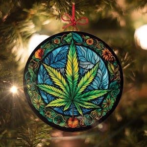 May include: A round Christmas ornament with a green marijuana leaf in the center. The leaf is surrounded by a colorful stained glass design with blue, green, yellow, and orange accents.