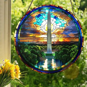 May include: A stained glass window depicting the Washington Monument in Washington, D.C. The monument is surrounded by trees and a reflecting pool. The sky is a vibrant blue with clouds and a setting sun. The stained glass is framed in a circular metal frame.