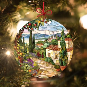 May include: A round stained glass ornament depicting a picturesque Italian countryside scene. The ornament features a stone path leading to a white house with a red tile roof, surrounded by lush greenery, cypress trees, and a distant mountain range. The sky is a vibrant blue with white clouds.
