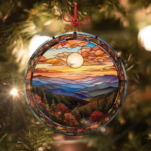 May include: A stained glass ornament depicting a sunset over a mountain range. The ornament features a bright orange sun, blue mountains, and a forest in the foreground. The ornament is round and has a red ribbon attached.