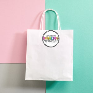 May include: White paper gift bag with a round sticker that says "We've been BUBBLING with excitement to meet you". The sticker has a black outline and colorful letters.
