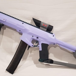 Könnte beinhalten: Eine Spielzeugpistole in Lavendelfarbe mit einem schwarzen Magazin und einem schwarzen Zielfernrohr. Die Pistole hat einen schwarzen Pistolengriff und einen grauen Schaft.