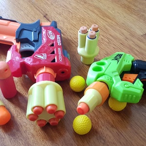 Puede incluir: Dos blasters de juguete, uno rojo y otro verde, con detalles naranjas y amarillos. El blaster rojo tiene una etiqueta "NERF MEGA EGGSHOCK". El blaster verde tiene una etiqueta "X-SHOT". Ambos blasters están rodeados de bolas de espuma amarillas.