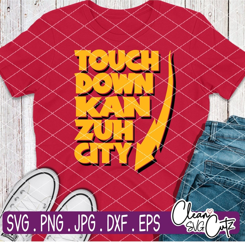 Op de afbeelding: Rood t-shirt met de tekst "TOUCH DOWN KAN ZUH CITY" in gele blokletters en een gele pijl. De afbeelding bevat een blauwe spijkerbroek en witte sneakers.