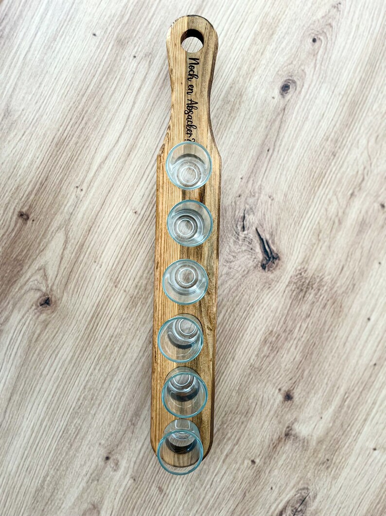 Pode incluir: Suporte de madeira para seis copos de shot. O suporte tem uma al&ccedil;a com o texto "Noch en Absacker?" gravado nele.