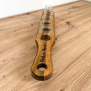 Pode incluir: Suporte de copos de shot de madeira com seis copos de shot e o texto "Noch ein Absacker?" gravado na al&ccedil;a.