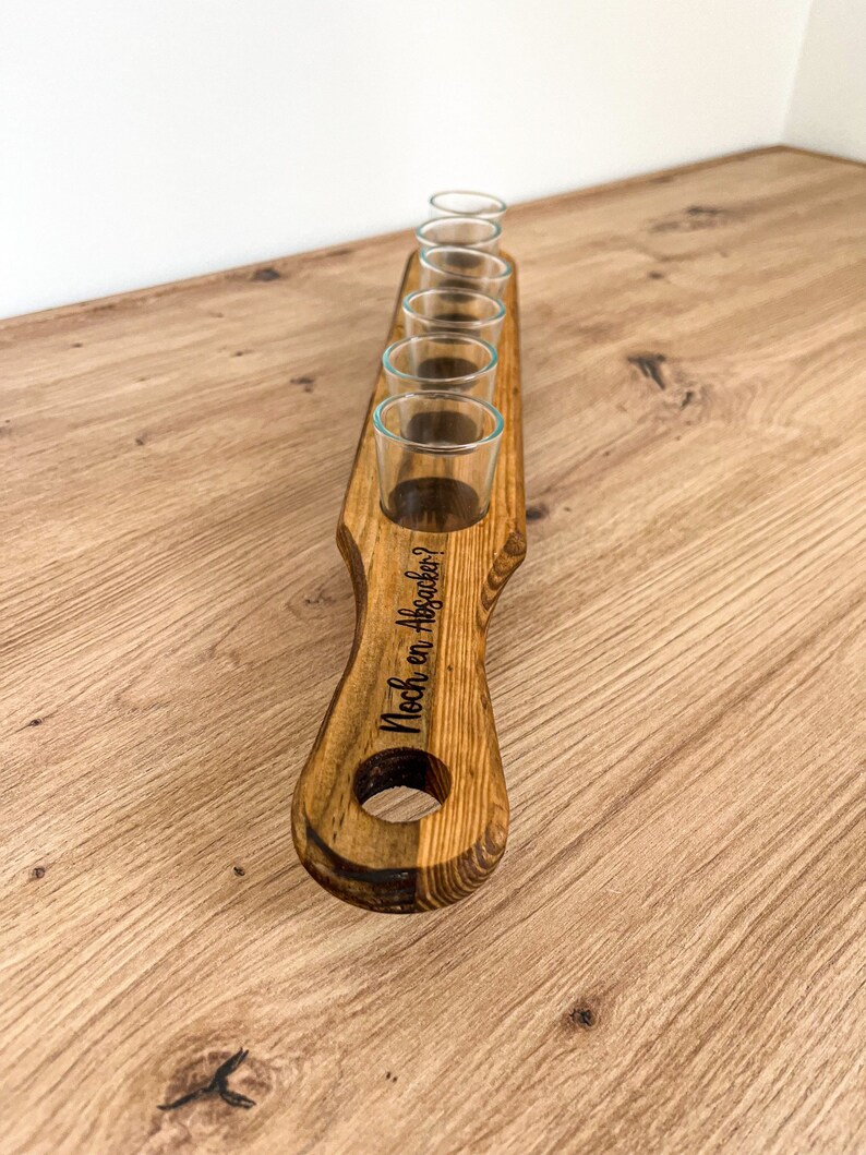 Pode incluir: Suporte de copos de shot de madeira com seis copos de shot e o texto "Noch ein Absacker?" gravado na al&ccedil;a.