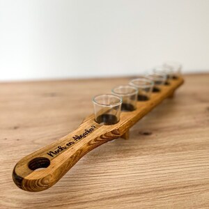 Pode incluir: Suporte de madeira para seis copos de shot. O suporte tem uma al&ccedil;a com o texto "Noch ein Absacker?" gravado nele.