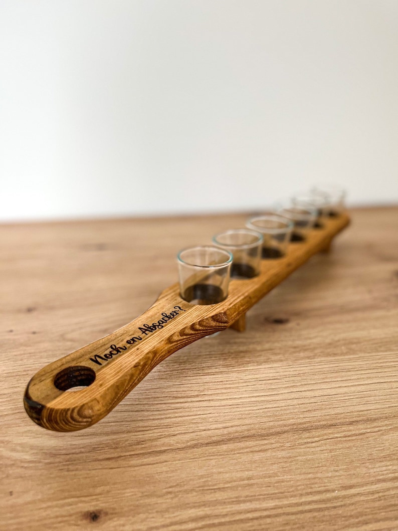 Pode incluir: Suporte de madeira para seis copos de shot. O suporte tem uma al&ccedil;a com o texto "Noch ein Absacker?" gravado nele.