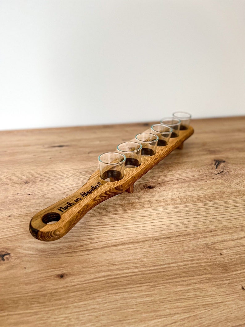 Pode incluir: Suporte de madeira para seis copos de shot. O suporte tem uma al&ccedil;a com o texto "Noch ein Absacker?" gravado nele.