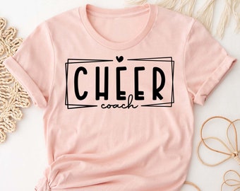 Peut inclure: Un t-shirt rose avec un texte noir qui dit "CHEER coach" dans un cadre noir avec un petit cœur au-dessus du texte.