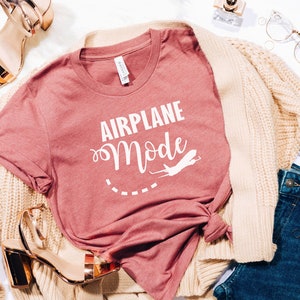 Puede incluir: Una camiseta rosa con el texto "Airplane Mode" en letras blancas. La camiseta se combina con una chaqueta de punto crema, unos vaqueros azules y tacones dorados.