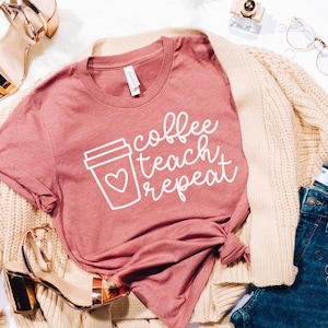 May include: A dusty rose t-shirt with the words "coffee teach repeat" in white script, featuring a coffee cup with a heart. The shirt is paired with blue jeans, a beige cardigan, and gold sandals.