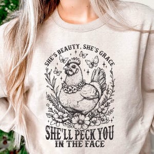 Może przedstawiać: Beżowa bluza z czarno-białą grafiką przedstawiającą kurę otoczoną kwiatami i motylami. Tekst brzmi: "She's Beauty, She's Grace, She'll Peck You in the Face."