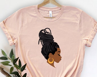 Op de afbeelding: Een lichtroze T-shirt met een afbeelding van een vrouw met een knot van dreadlocks en gouden oorringen.