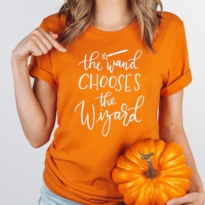 May include: A woman wearing an orange t-shirt with the text "the wand chooses the wizard" in white lettering. She is holding a small orange pumpkin.