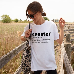 以下が含まれることがあります： 黒い文字で「seester [se-ster] - 名詞 姉妹のようなもの、ただもっとクール。参照: 一生の親友」と書かれた白いTシャツ