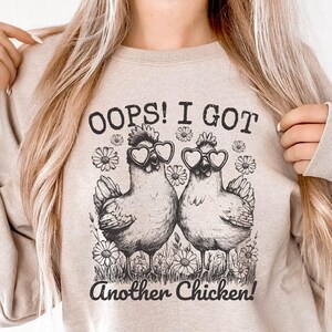 Puede incluir: Una sudadera beige con un gráfico negro de dos pollos con gafas de sol en forma de corazón y el texto "OOPS! I GOT Another Chicken!"