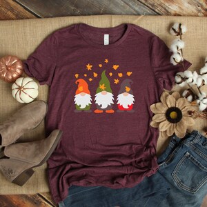 May include: A maroon t-shirt with a graphic of three gnomes wearing hats and standing in a row. The gnomes are surrounded by small orange leaves.