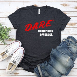 Puede incluir: Una camiseta gris oscuro jaspeada con las palabras "DARE. TO KEEP KIDS OFF DRUGS." impresas en blanco. La palabra "DARE" está en rojo y estilizada.
