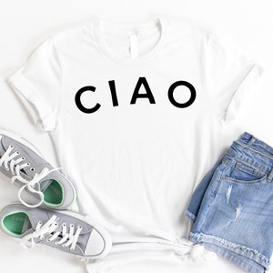 May include: White t-shirt with the word "CIAO" printed in black on the front. The shirt is laid flat on a white surface with a pair of gray sneakers and blue denim shorts.