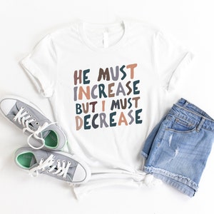 K&ouml;nnte beinhalten: Wei&szlig;es T-Shirt mit dem Text "HE MUST INCREASE BUT I MUST DECREASE" in bunten, verspielten Buchstaben. Das Shirt wird mit Jeans-Shorts und grauen Sneakern kombiniert.