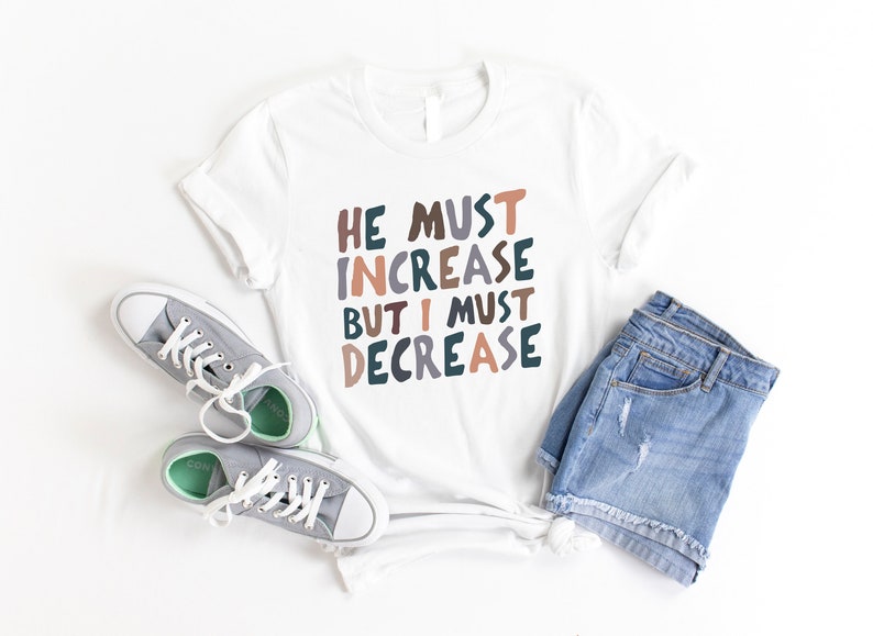 K&ouml;nnte beinhalten: Wei&szlig;es T-Shirt mit dem Text "HE MUST INCREASE BUT I MUST DECREASE" in bunten, verspielten Buchstaben. Das Shirt wird mit Jeans-Shorts und grauen Sneakern kombiniert.