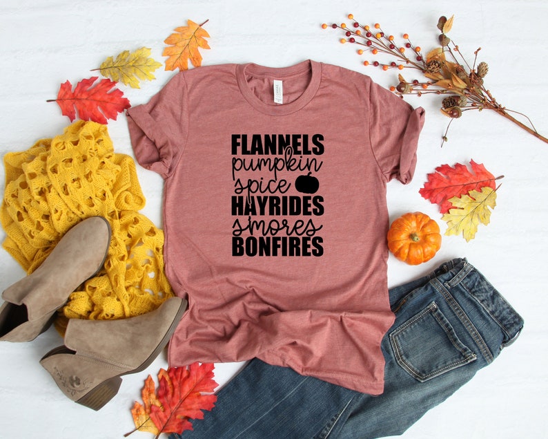 May include: A maroon t-shirt with the text "Flannels, pumpkin spice, hayrides, smores, bonfires" in black. The shirt is laid out on a white surface with autumnal decorations.