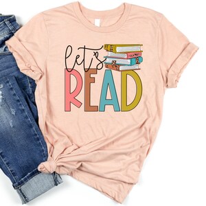 May include: A pink t-shirt with the text "Let's Read" in colorful letters. A stack of books with colorful covers is above the text.