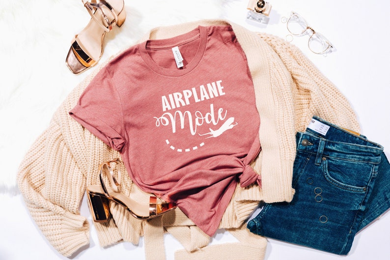 Puede incluir: Una camiseta rosa con el texto "Airplane Mode" en letras blancas. La camiseta se combina con una chaqueta de punto crema, unos vaqueros azules y tacones dorados.