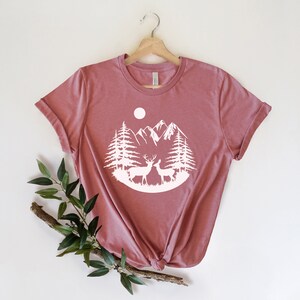 Puede incluir: Camiseta de color malva con un gr&aacute;fico blanco de una escena de monta&ntilde;a con dos ciervos, &aacute;rboles y una luna. La camiseta se muestra en una percha de madera sobre un fondo blanco. Una ramita de vegetaci&oacute;n se coloca en la parte inferior izquierda.