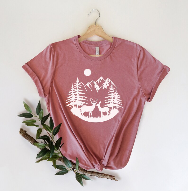 Puede incluir: Camiseta de color malva con un gr&aacute;fico blanco de una escena de monta&ntilde;a con dos ciervos, &aacute;rboles y una luna. La camiseta se muestra en una percha de madera sobre un fondo blanco. Una ramita de vegetaci&oacute;n se coloca en la parte inferior izquierda.