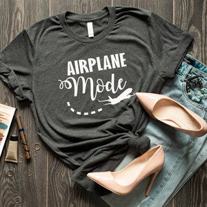 Puede incluir: Una camiseta gris oscuro con el texto "Airplane Mode" en letras blancas y un gr&aacute;fico de avi&oacute;n blanco. La camiseta est&aacute; colocada sobre una superficie de madera con un par de tacones altos de color rosa claro y unos vaqueros azules.