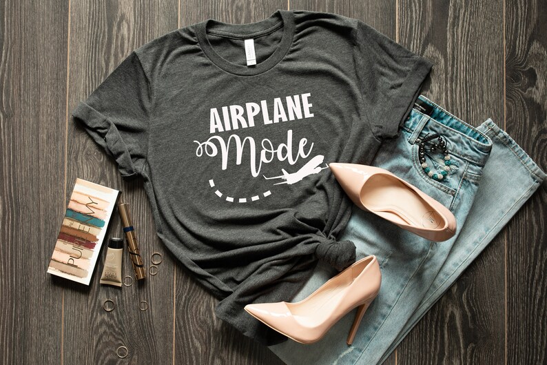 Puede incluir: Una camiseta gris oscuro con el texto "Airplane Mode" en letras blancas y un gr&aacute;fico de avi&oacute;n blanco. La camiseta est&aacute; colocada sobre una superficie de madera con un par de tacones altos de color rosa claro y unos vaqueros azules.
