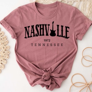 May include: A mauve colored t-shirt with a black graphic that says "NASHVILLE 1972 TENNESSEE" with a guitar silhouette.