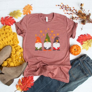 May include: A maroon t-shirt with a graphic of three gnomes wearing hats and holding pumpkins. The gnomes are surrounded by falling leaves.