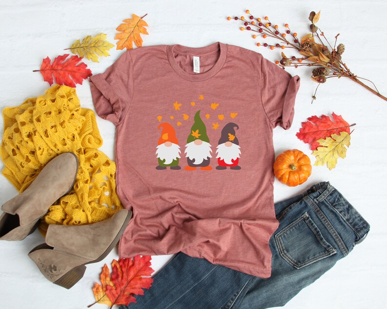 May include: A maroon t-shirt with a graphic of three gnomes wearing hats and holding pumpkins. The gnomes are surrounded by falling leaves.
