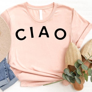 May include: A light pink t-shirt with the word "CIAO" printed in black on the front. The shirt is laying on a white surface with a straw hat and sandals.