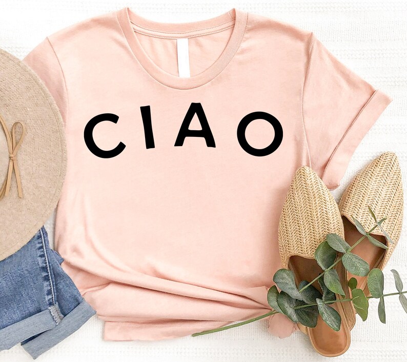 May include: A light pink t-shirt with the word "CIAO" printed in black on the front. The shirt is laying on a white surface with a straw hat and sandals.