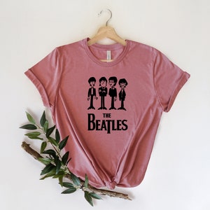 Puede incluir: Una camiseta color malva con un gr&aacute;fico negro de los Beatles. El gr&aacute;fico presenta una imagen de dibujos animados de los miembros de la banda y el texto "THE BEATLES".