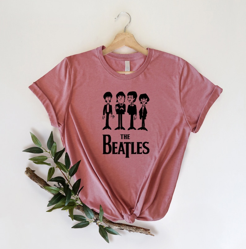 Puede incluir: Una camiseta color malva con un gr&aacute;fico negro de los Beatles. El gr&aacute;fico presenta una imagen de dibujos animados de los miembros de la banda y el texto "THE BEATLES".