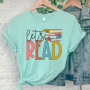 May include: A light blue t-shirt with the text "let's read" in colorful letters. A stack of books with colorful covers is behind the text.