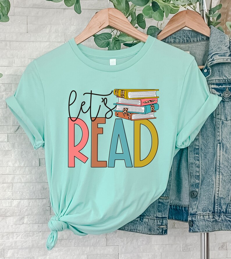 May include: A light blue t-shirt with the text "let's read" in colorful letters. A stack of books with colorful covers is behind the text.